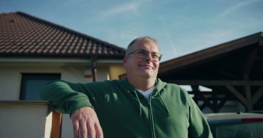 Evin önündeki konut kapısına yaslanan adam, gülümseyen ve rahat, banliyö yaşam tarzı ve mülk çevresi odaklı