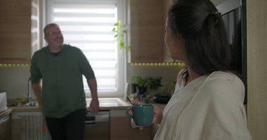 İki yetişkin modern bir mutfakta neşeli bir şekilde sohbet ediyorlar. Mavi bir kupa tutan ve gülümseyen bir kadın. Tezgaha yaslanan bir adam, rahat bir atmosfer.