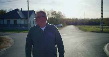 Huzurlu bir banliyö sokağında yan bakan orta yaşlı bir adam, gözlük takıyor ve sakin bir mahallede yansıtıcı ve düşünceli bir tavır sergiliyor.