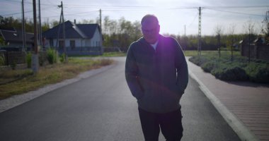 Orta yaşlı bir adam, sakin bir sokakta elleri ceplerinde, sakin bir duruş ve sakin bir mahalle atmosferi sergiliyor.