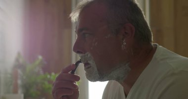 Jiletle tıraş olan, başını hafifçe eğen, temiz bir tıraş için baskı uygulayan, yüzünde köpük olan, arka planda banyo detayları, hijyen ve bakım yapan bir adam.