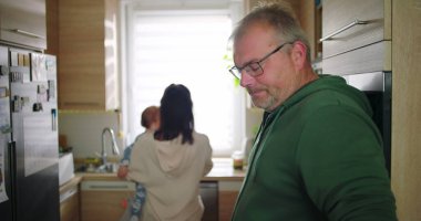 Adam elinde fincanla gülümserken, anne mutfakta bebeği tutuyor, neşeli aile anları günlük ev rutinlerinde, gündelik ve sıcak etkileşimler