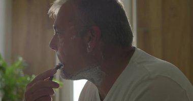 Jiletle tıraş olan, başını hafifçe eğen, temiz bir tıraş için baskı uygulayan, yüzünde köpük olan, arka planda banyo detayları, hijyen ve bakım yapan bir adam.