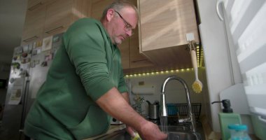 Bir adam mutfak lavabosundaki musluk suyunun altında mavi bir tabağı duruluyor, günlük ev işleri sırasında bulaşık takımlarını temizlemeye odaklanıyor.