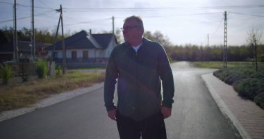 Orta yaşlı bir adam, sakin bir sokakta elleri ceplerinde, sakin bir duruş ve sakin bir mahalle atmosferi sergiliyor.