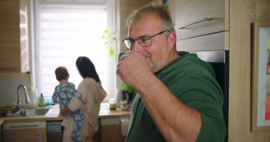 Adam elinde fincanla gülümserken, anne mutfakta bebeği tutuyor, neşeli aile anları günlük ev rutinlerinde, gündelik ve sıcak etkileşimler