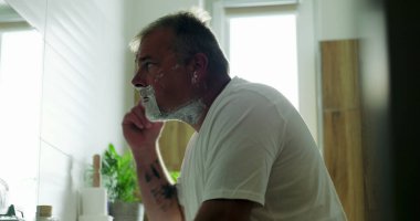 Jiletle tıraş olan, çene açısını hassasiyete göre ayarlayan, yüzdeki köpüğü tıraş eden düşünceli ifade, banyo arka planı, sabah bakım rutini.