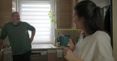 İki yetişkin modern bir mutfakta neşeli bir şekilde sohbet ediyorlar. Mavi bir kupa tutan ve gülümseyen bir kadın. Tezgaha yaslanan bir adam, rahat bir atmosfer.