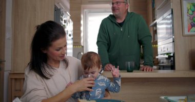 Anne, bebeğin yemek masasında otururken su içmesine yardım ediyor, baba arka planda dikiliyor, sıcak bir aile mutfağında.