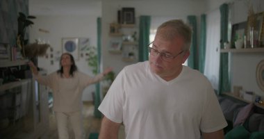 Erkek, arka plandaki kadınla tartışırken gözle görülür şekilde üzgün, oturma odasında ilişki gerilimi, duygusal ve dramatik anlaşmazlık anı