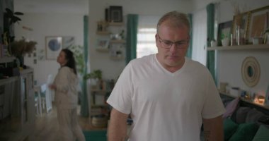 Erkek, arka plandaki kadınla tartışırken gözle görülür şekilde üzgün, oturma odasında ilişki gerilimi, duygusal ve dramatik anlaşmazlık anı