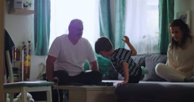 Aile rahat bir anı paylaşıyor, baba masaya oturuyor, genç çocuk öne eğiliyor, ve anne rahat oturma odasında kanepeye odaklanıyor.