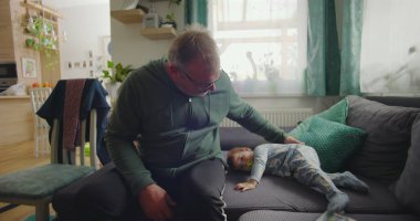 Babam kanepeye oturmuş bebeği nazikçe rahatlatıyor. Emzik ile uzanıyor, rahat bir aile salonunda. Hafif ışıklandırması ve sakin bir atmosferi var.