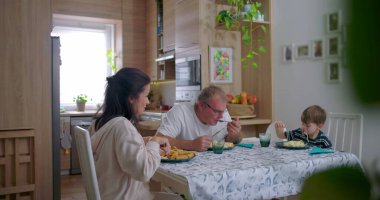 Aile masada sakin bir yemeği paylaşıyor, ebeveynler ve çocuklar arasındaki gerçek etkileşim, sıcak bir aile ortamının altını çiziyor.