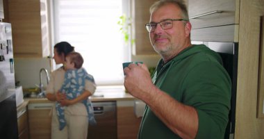 Anne bebeğini mutfakta tutarken, adam gülümsüyor ve ön planda bardağı tutuyor. Neşeli aile etkileşimi günlük sabah rutini, ev hayatı.