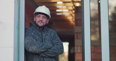 İnşaat işçisi kapıda kolları çaprazlanmış, düşünceli bir şekilde dik dik bakıyor, başlık takıyor, arka plan yapım aşamasında olduğunu gösteriyor, kararlılığı ve odaklanmayı simgeliyor.