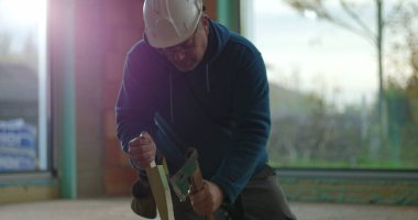 İnşaat işçisi odun parçalarını odaklama ve kuvvetle çekiçliyor, iç inşaat sahasında diz çöküyor, malzemeleri güvence altına alıyor, inşaat projesi devam ediyor.