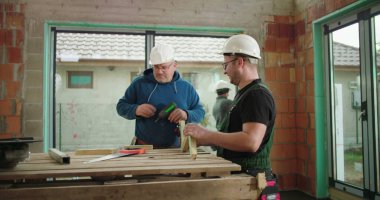 İnşaat işçileri inşaat alanındaki ahşap parçaları inceliyor ve hizalıyorlar, ikisi de kask takıyor, bina yapısında takım çalışması yapıyor, hassasiyet ve güvenliğe odaklanıyor.