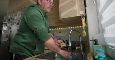 Adam mutfak lavabosunda bulaşık yıkıyor, akmakta olan bir kâseyi duruluyor, modern bir evde ev temizliği yapıyor.