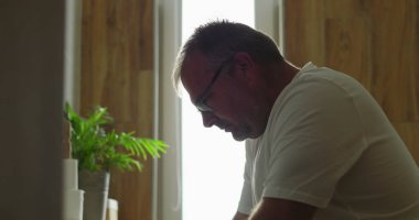 Banyo aynasının önündeki profil görünümlü adam, nazik bir ifadeyle, içeriden yansıyan, arka pencereden süzülen ışık, sakin ve düşünceli bir an.