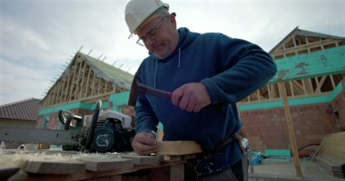 İnşaat işçisi tahta bloğa çivi çakıyor, tezgahın yanında elektrikli testere, arka planda bina yapısı, ustalık ve inşaat alanında kendini adamışlık.