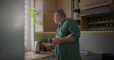 Adam mavi bardağa sıcak su dolduruyor, mutfakta çay ya da kahve hazırlıyor, sabah rutini, günlük ev hayatı, doğal ışık alan modern mutfak.