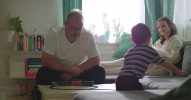 Baba sehpada genç bir çocukla oyuncak tren oyunu oynuyor, anne kanepede küçük beyaz bir köpekle gülümsüyor, neşeli bir aile, sıcak bir evde oturma odasında kaynaşıyor.
