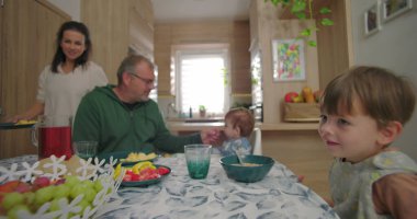 Aile sıcak bir mutfakta birlikte yemek yiyor, ebeveynler yemek yerken çocuklarla etkileşime geçiyor, yemek sırasında bağ ve birlikteliği vurguluyor.
