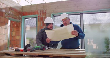 Bitmemiş binadaki ahşap masanın detaylı planlarını inceleyen inşaat işçileri, projeyi tartışıyorlar, güvenlik kaskı takıyorlar, takım çalışması yapıyorlar, işbirliği yapıyorlar.