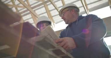 İnşaat danışmanı ve iş arkadaşı proje detaylarını, takım çalışmasını ve rehberliği tartışıyorlar. Her ikisi de arka planda yapısal çerçeveye sahip güvenlik miğferlerinde.