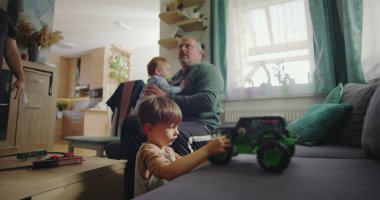 Baba iki çocukla oturuyor, biri yeşil bir oyuncak kamyonla oynuyor, diğeri de sıcak bir aile salonunda, modern ve sıcak bir dekorla izliyor.