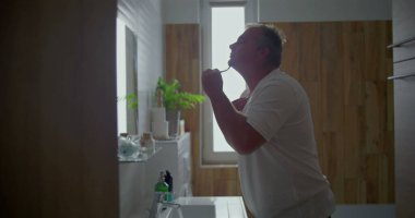 Adam banyoda tıraş oluyor, başını arkaya eğip çene altını tıraş ediyor, köpük uyguluyor, doğal banyo unsurları görülüyor, hijyen ve kişisel bakım üzerine odaklanıyor.