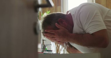 Adam banyo lavabosunda yüzünü suyla yıkıyor, elleri kavrulmuş ve su akıyor, kişisel hijyene odaklanıyor ve temiz ev ortamında ferahlatıcı sabah rutinine odaklanıyor.