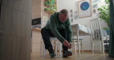 Ahşap mobilyalar ve sıcak ev dekorlarıyla çevrili, rahat bir aile yemek odasında çömelmiş ve botlarını bağlayan bir adam.