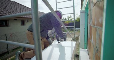 Yalıtım tahtasına yapıştırıcı süren inşaat işçisi, duvar kurulumu, güvenlik kaskı, arka planda merdiven ve yapı iskelesi olan dış yapı projesi için hazırlık yapıyor.