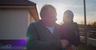 Father holding baby with pacifier in backyard, gently adjusting child's outfit, showcasing nurturing bond and love in a serene family environment