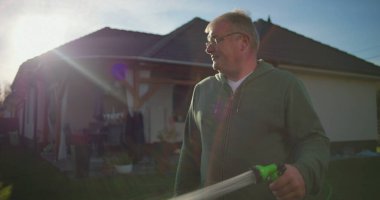 Bahçe hortumunu arka bahçeyi sulamak için kullanan bir adam varoş ortamında çimlere doğru kıvrılan su akıntısı arka planda görünen ev ve veranda, sakin bir an.