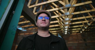 İnşaat işçisi açık çatı altında duruyor, başlık ve gözlük takıyor, inşaatın ilk aşamasında inşaat projesine odaklanıyor ve kararlılıkla bakıyor.