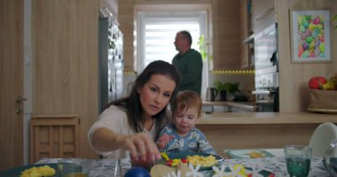 Anne ve bebek kahvaltı masasında, neşeli bebekle beslenme anını paylaşıyorlar, aile mutfakta basit anların tadını çıkarıyor, paylaşılan yemek sırasında gelişigüzel bağ kuruyorlar.