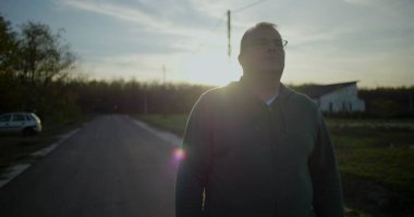 Gün batımında dışarıda yürüyen gözlüklü orta yaşlı bir adam, rahat ve düşünceli bir yüz ifadesi, kırsal alan, akşam ışığı, sakin atmosfer, kırsal huzur.