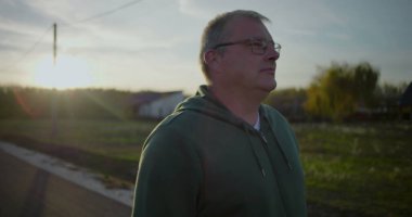 Gün batımında dışarıda yürüyen gözlüklü orta yaşlı bir adam, rahat ve düşünceli bir yüz ifadesi, kırsal alan, akşam ışığı, sakin atmosfer, kırsal huzur.