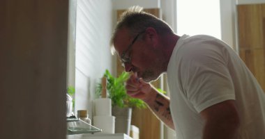 Banyo lavabosunda dişlerini fırçalayan adam, günlük sabah rutininin bir parçası olarak diş hijyenine odaklanıyor, minimalist ve temiz ortamlarda pencereden ışık yayılıyor.