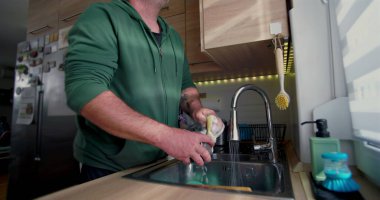 Adam mutfak lavabosunun altında sabun ve süngerle bulaşık yıkıyor, rahat bir ev mutfağında temizlik ve günlük ev işlerine odaklanıyor.