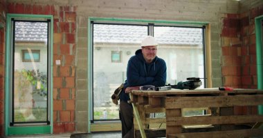 İnşaat işçisi kısmen inşa edilmiş bir yapıda, kollarını kavuşturmuş, kaskını takmış, inşaat alanındaki görevine odaklanmış.