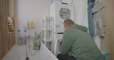 Çamaşırları yıkama makinesiyle küçük banyoda düzenleyen, raflarla, havlularla ve banyo aksesuarlarıyla çevrili, günlük ev işlerini vurgulayan bir adam.