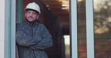 İnşaat işçisi kapıda kolları çaprazlanmış, düşünceli bir şekilde dik dik bakıyor, başlık takıyor, arka plan yapım aşamasında olduğunu gösteriyor, kararlılığı ve odaklanmayı simgeliyor.