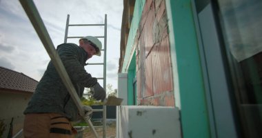 Dış duvardaki yalıtım panelini malayla temizliyorum, inşaat işçisi, tuğla inşaatında çalışıyorum, miğfer ve eldivenle merdiven üzerinde duvar işçiliği yapıyorum.