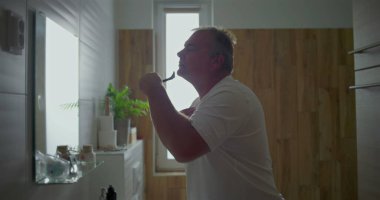 Adam banyoda tıraş oluyor, başını arkaya eğip çene altını tıraş ediyor, köpük uyguluyor, doğal banyo unsurları görülüyor, hijyen ve kişisel bakım üzerine odaklanıyor.