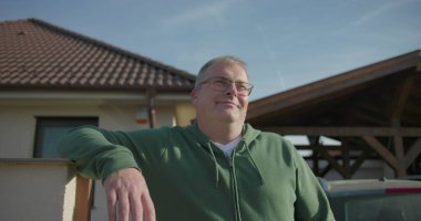 Evin önündeki iskan kapısına yaslanan bir adam, garaj yolu ve park edilmiş arabanın yanında gelişigüzel poz veriyor, banliyö ortamındaki rahat açık hava anına odaklanıyor.