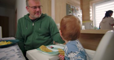 Baba, ufaklığın waffle ve meyveyle kahvaltı yapmasını seyrediyor. Sıcak ve samimi bir anı paylaşıyor. Aile sabahı rutininde, sıcak bir ev mutfağında.